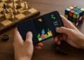 Pessoa jogando Tetris no celular ao lado de tabuleiro de xadrez e cubo mágico, representando jogos clássicos.
